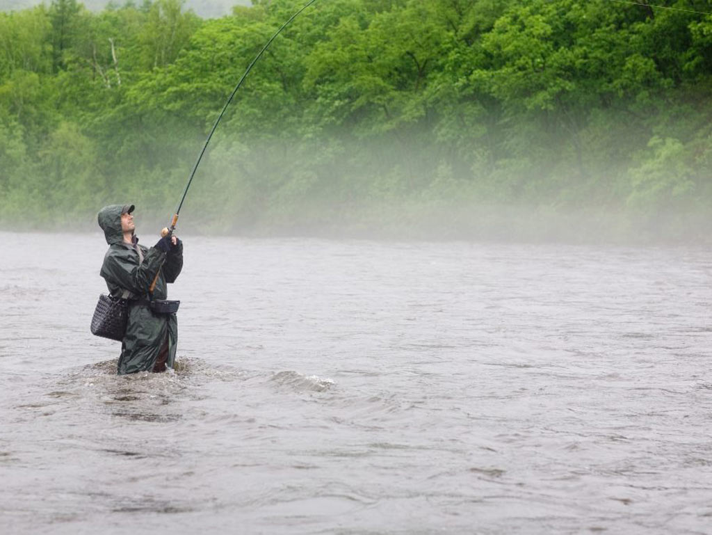 A view across a river towards an angler fishing in a river in the rain, while wearing full fishing gear