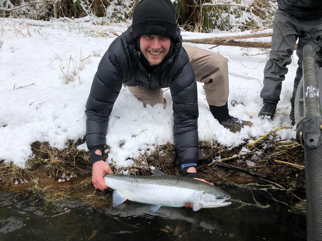 An angler leaning towards water on a river bank, holding a winter Steelhead he caught slightly above water, with snow covering the shoreline.
