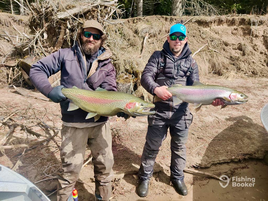 Two men in baseball caps and sungalsses stand on a shore holding a large Rainbow Trout each on a sunny day