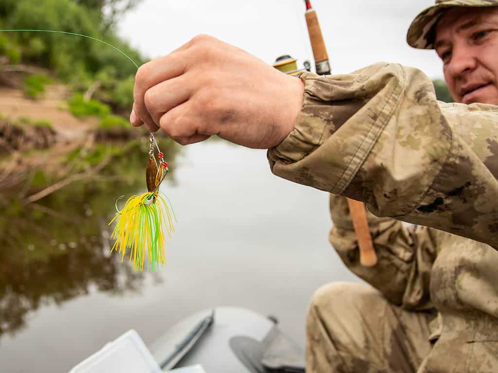 A closeup image of a spinnerbait being used as lure on the end of a fishing line being held by a male angler on a boat on a cloudy day