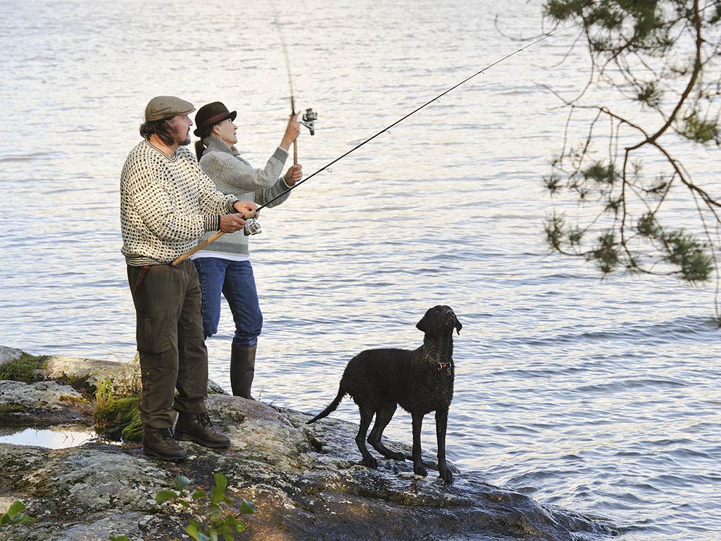 A couple fishing with a spinning rod and reel combo, as the woman attempts to retrieve a catch, with a black dog next to them looking on