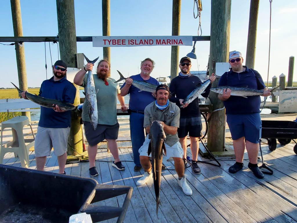 A group photo of six anglers posing with their fish caught during a deep sea fishing trip on the wooden dock of Tybee Island Marina