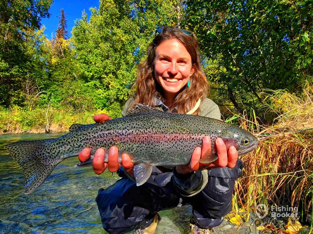 A woman smiling while holding a Rainbow Trout next to a river and surrounded by greenery on a clear day