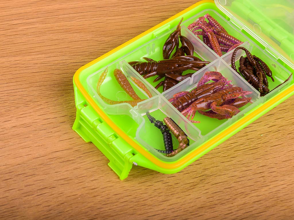 A closeup of a fluorescent tackle box sitting on a wooden desk with a number of colorful soft plastic lures in it