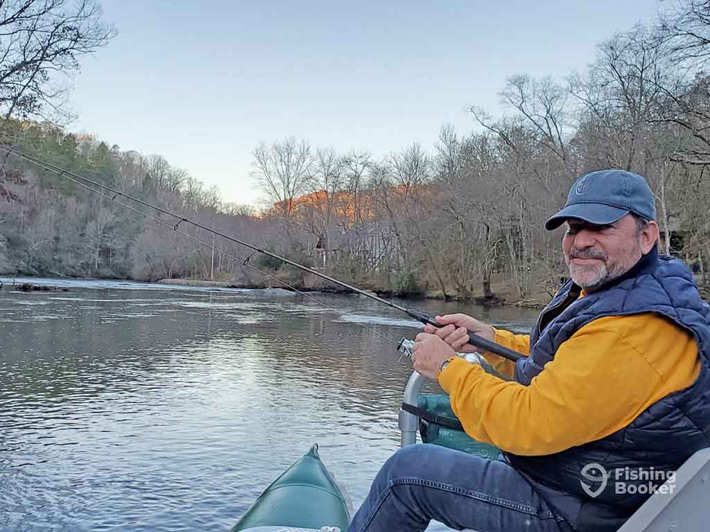 A man in a yellow sweater and navy gillet sitting on a fishing boat in North Carolina and casting his line for Trout on a clear but crispy day