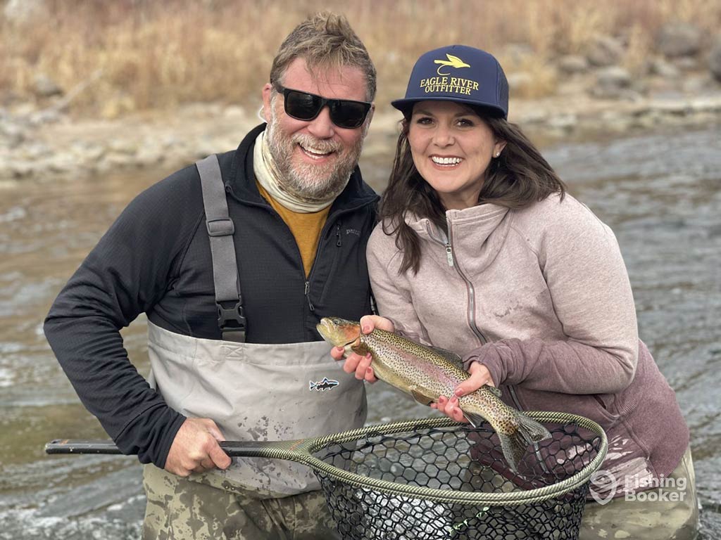 A man and a woman posing on a fishing trip in Colorado, as the man holds a fishing net and the woman a Rainbow Trout on a grey day