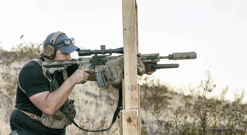 Photo of a man bracing a firearm against a wooden pole.