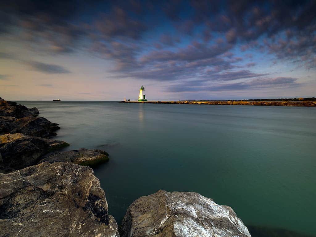 A view across the water at sunset from a rocky shoreline towards Port Dalhousie lighthouse in St.Catharines, with clouds visible overhead