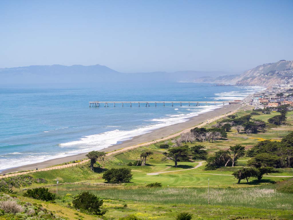 A view from a hill looking towards the beachfront of Pacifica, California, with the city's famed fishing pier visible as it juts out into the ocean.