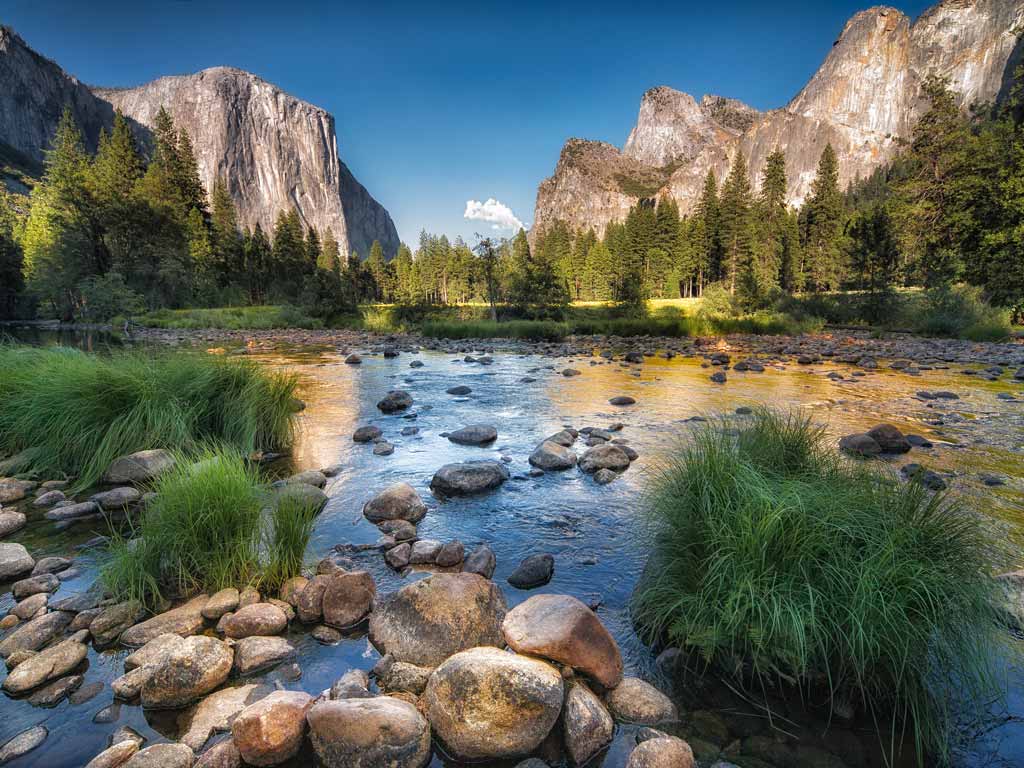 A spectacular photo of the Yosemite National Park rock formations viewed from a shallow river's shore on a clear day. 