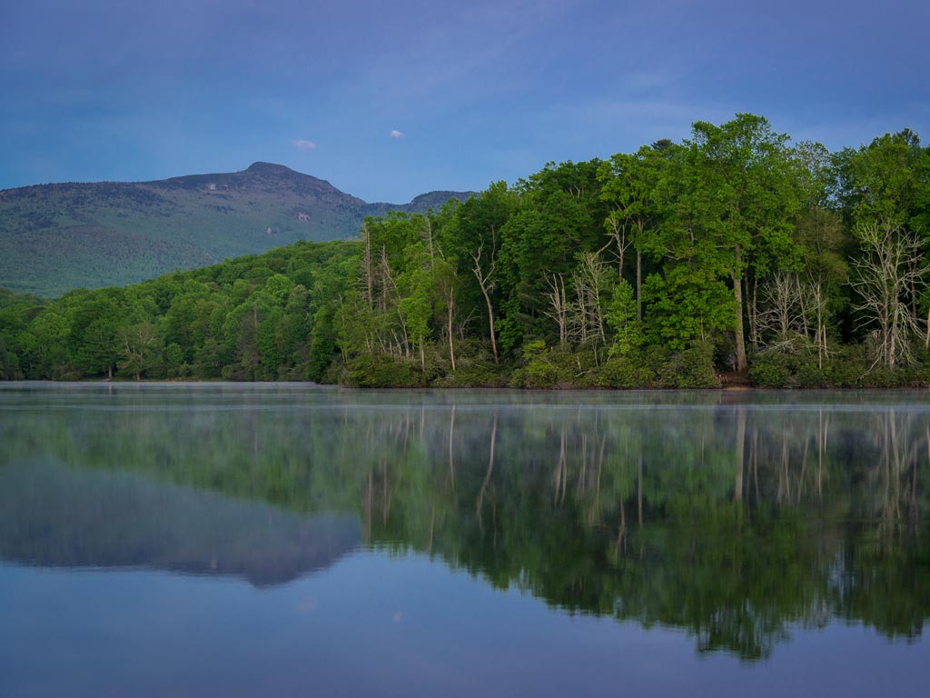 A scenic view of Julian Price Lake near Blowing Rock, NC, the lake's waters are calm and the shoreline forest trees are reflecting off the surface in a beautiful way.