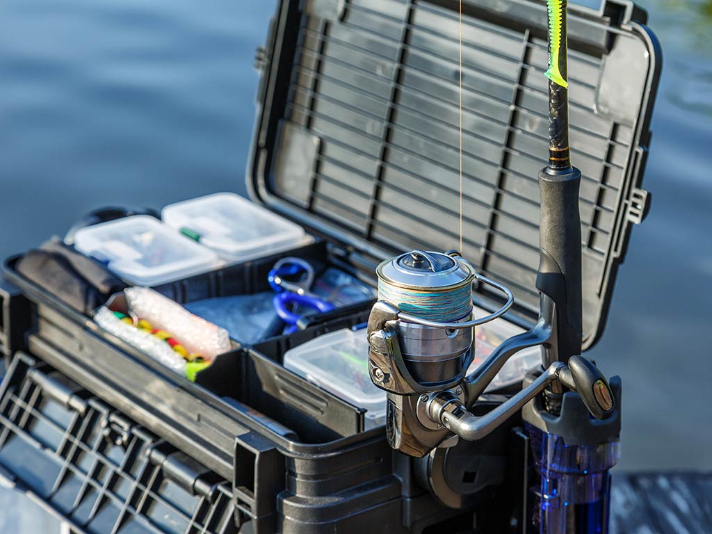 A large tackle box full of gear with a fishing rod and reel stood upright next to it, with the water out of focus behind it