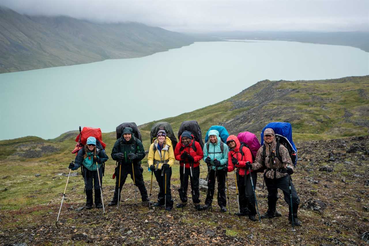 A group of smiling hikers standing in front of a lake in Alaska. It's raining and they're wearing rain jackets.