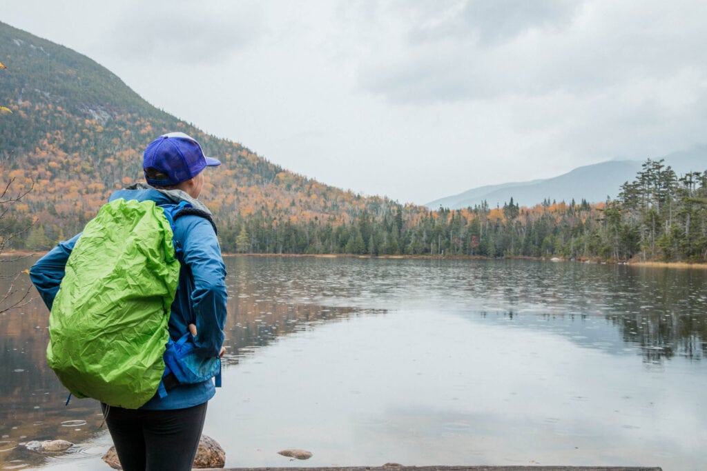woman standing next to a lake with fall foliage covering the hillside. She is wearing rain gear and a rain cover over her hiking backpack