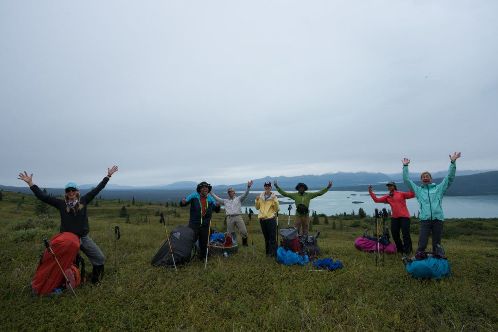 A group of backpackers smile at the camera with their packs on the ground in Alaska on a cloudy, rainy day.