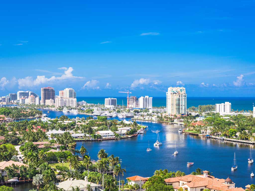 An aerial image of Fort Lauderdale on a sunny day and the Intracoastal Waterway that flows through it, the channel is visible in the forefront while buildings and the ocean are visible in the distance.
