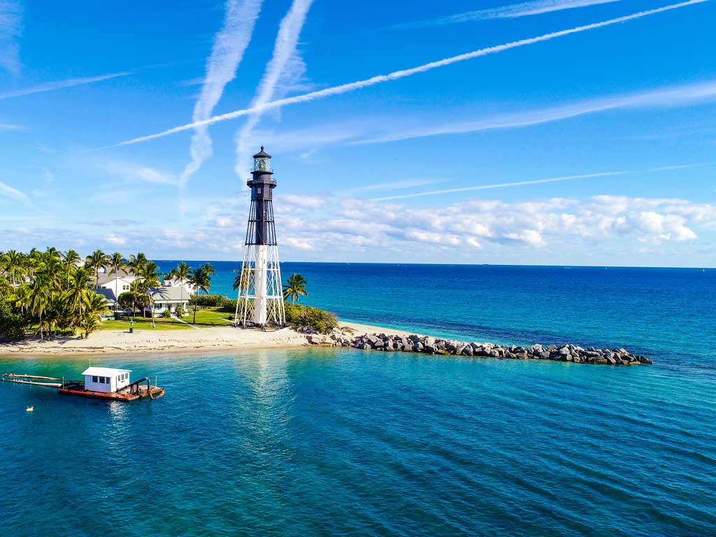 A photo of the Hillsboro Inlet Lighthouse on a sunny day, with a boat passing by the lighthouse and the deep blue ocean waters surrounding the jetty stretching from the land.
