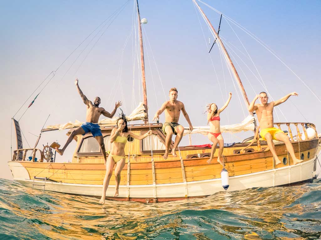 A group of multiracial people, three men and two women, jumping into the water from a sailboat. The photo was taken from the water with a slight fisheye effect.