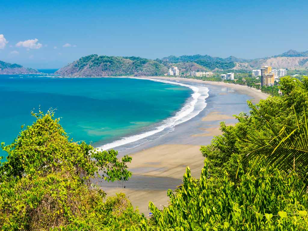 A photo of Jaco Beach in Costa Rica taken from distance between two shrubs, with the beautiful turquoise waters visible as the beach curves in a crescent shape.