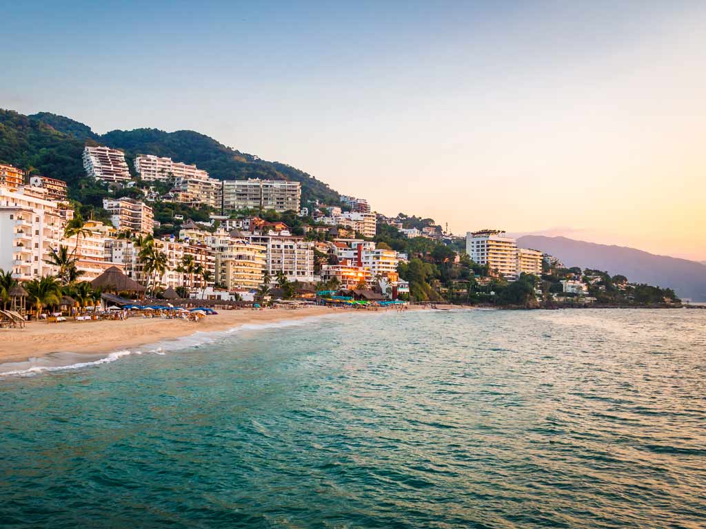 A sunset shot of the Puerto Vallarta coastline, with the city visible to the left of the photo as well as the Sierra Madre mountains behind it.