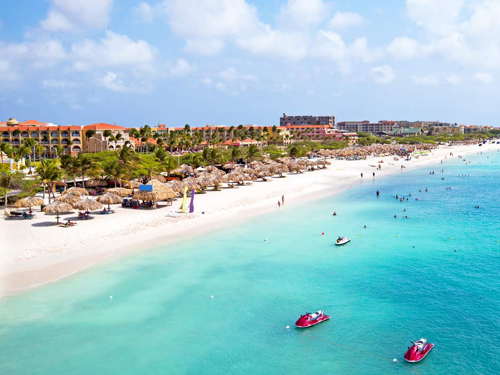 An aerial shot of the Eagle Beach in Aruba, with white sands and aqua blue waters creating a stunning scenery, while the local buildings are visible in the background.