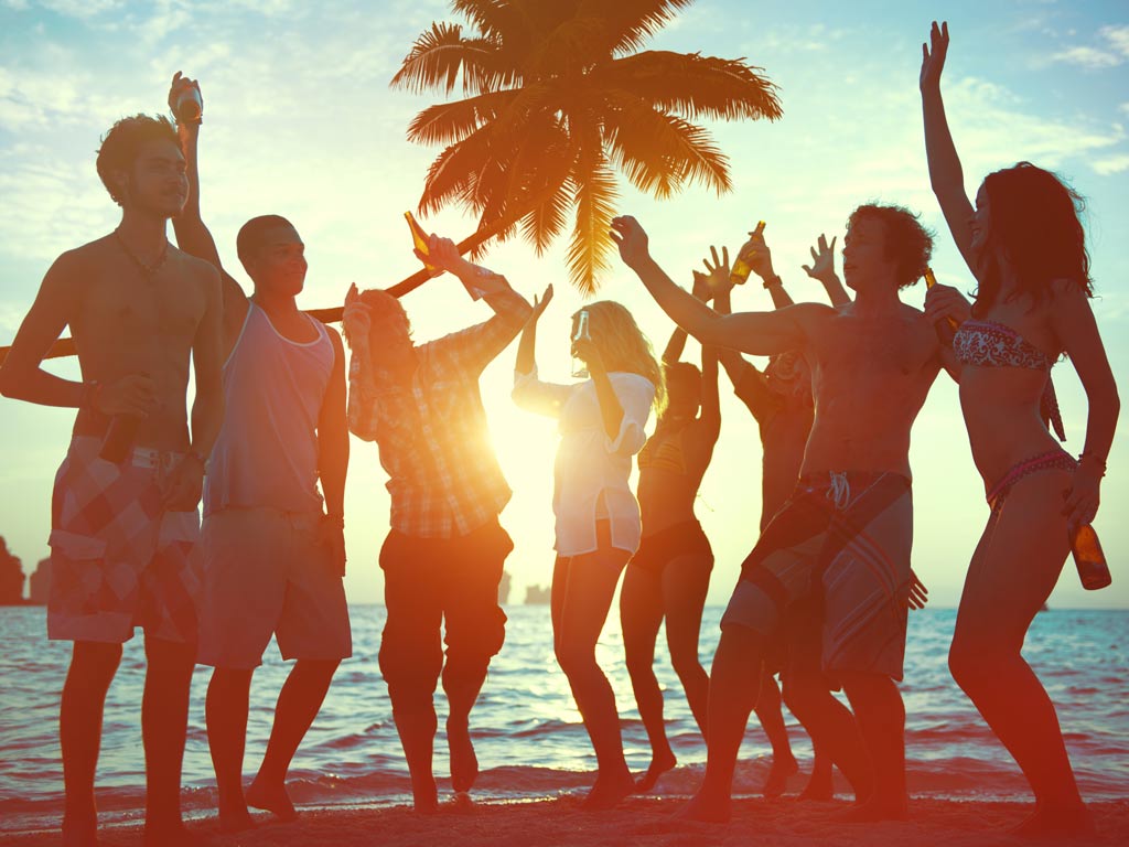 A photo of a group of people dancing on a beach at sunset, with the camera pointing at the setting sun and the sunlight partially obscuring the men and women.