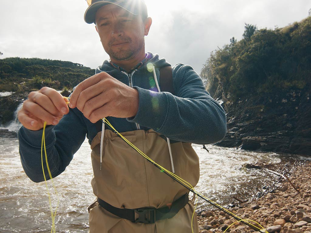 A man wading waist-deep in the waters of a river, tying a knot on the end of his fly fishing line on a bright day