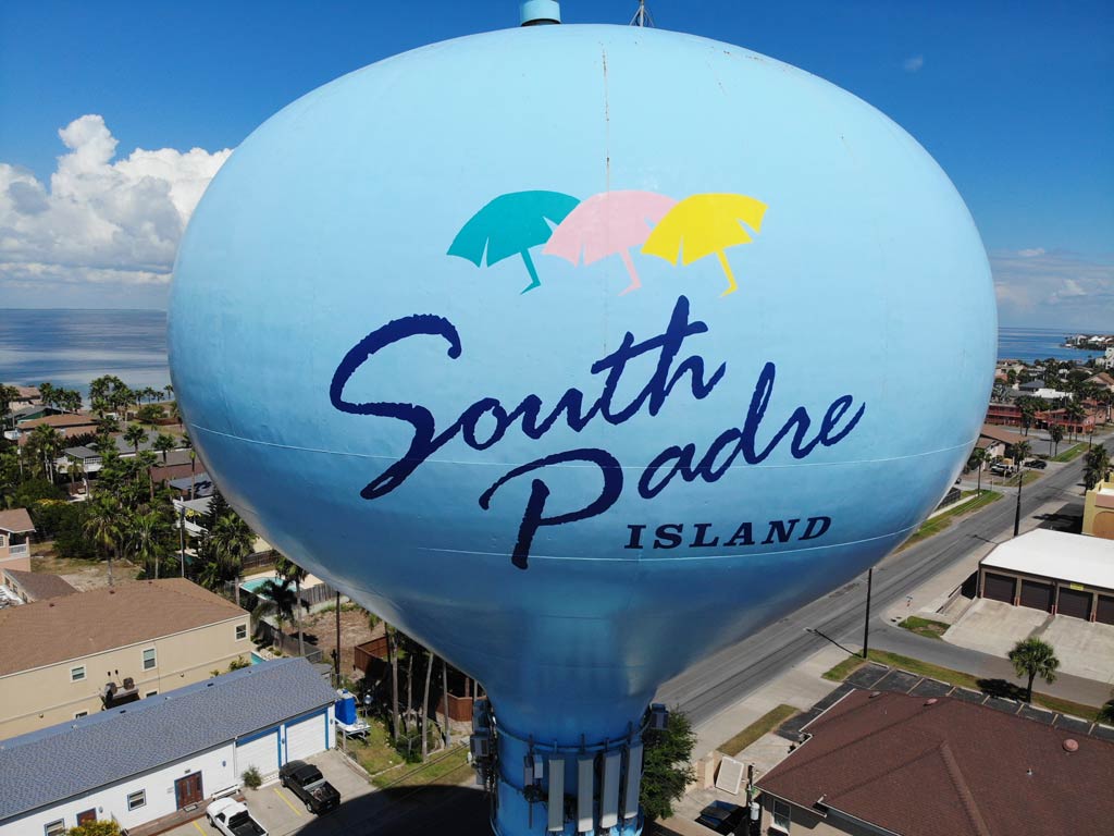 A view of a big oval blue South Padre Island sign as seen from above and with the ocean, streets, and houses visible in the background