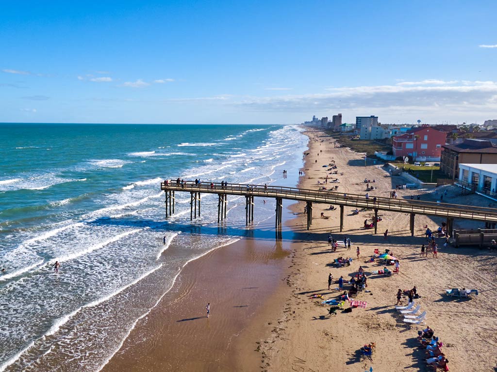 An aerial view from the side of South Padre Island's wooden pier, with the wavy seas, houses, and sandy beach dotted with beachgoers visible on a clear day