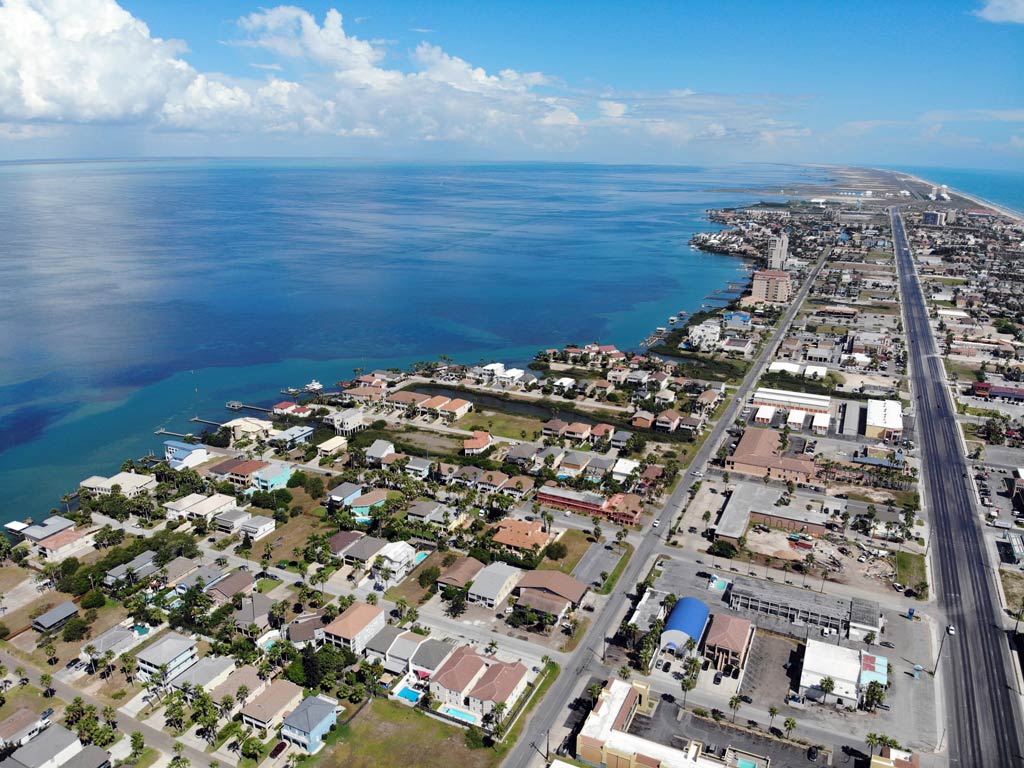 An aerial photo of South Padre Island featuring the ocean, streets, houses, and greenery on a bright and sunny day