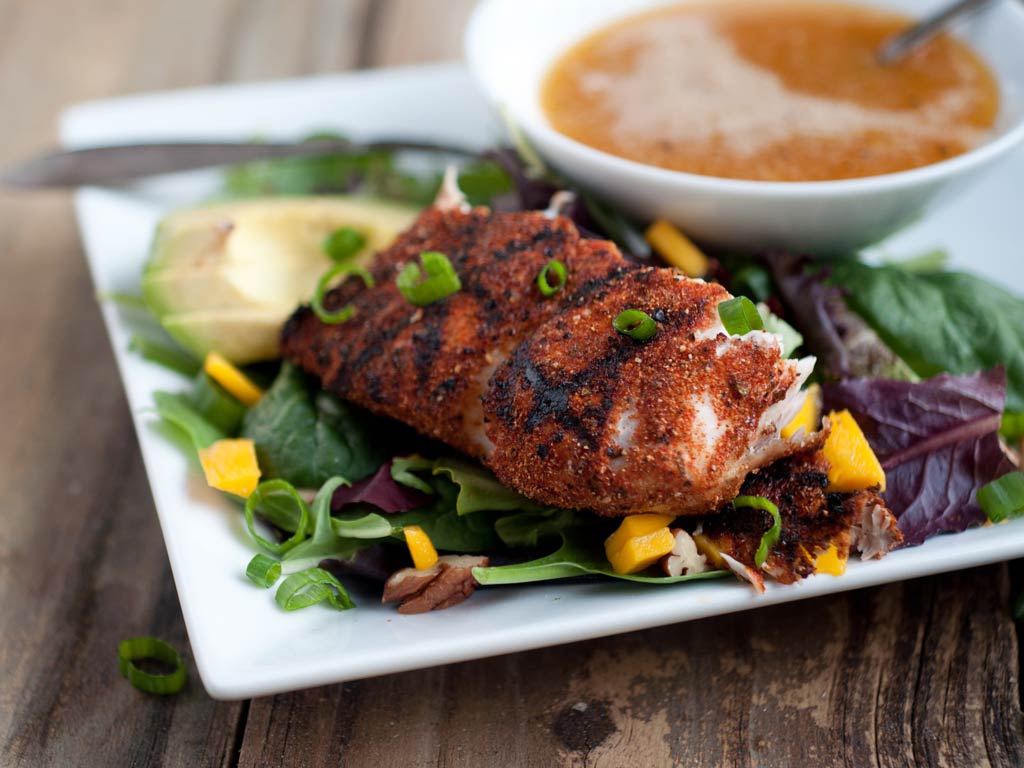 A photo of a grilled and blackened fish fillet served on a white plate along with salad, avocado, mango, and a citrus sauce