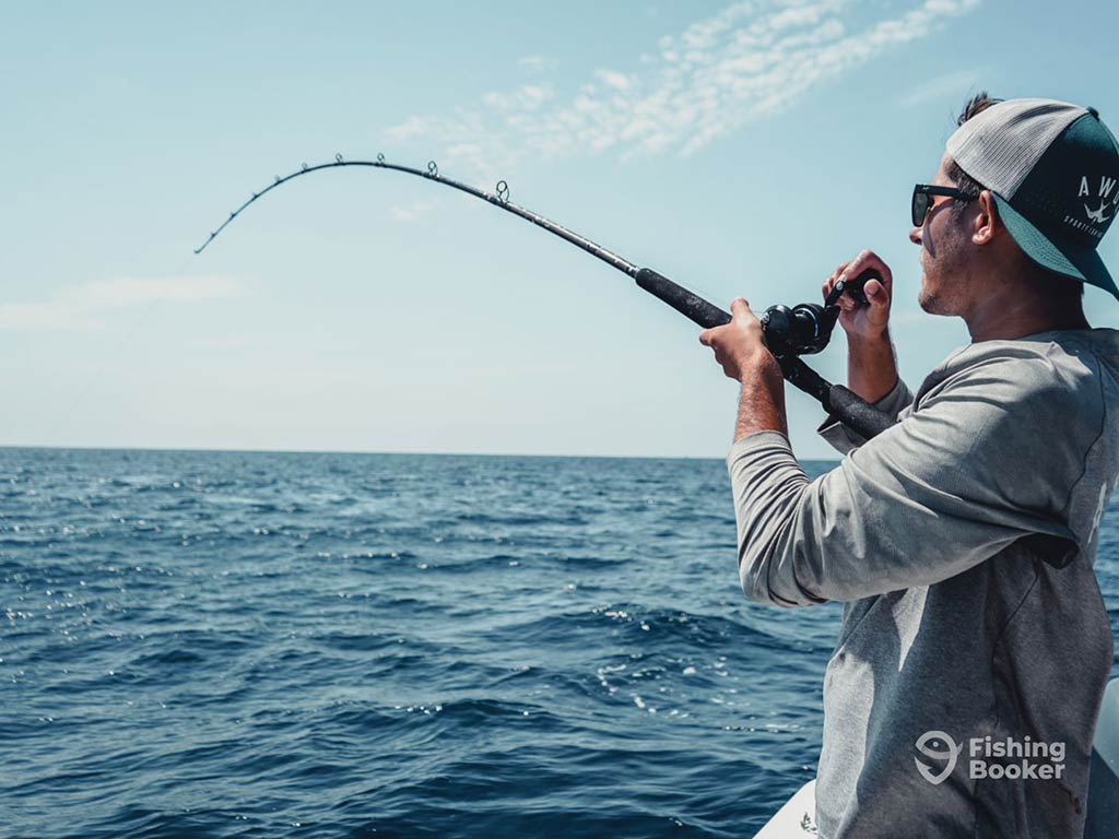 An angler in a backwards baseball cap holds a fishing rod up to his chest and attempts to reel in whatever is on the end of his line in the water that's visible on the left of the image