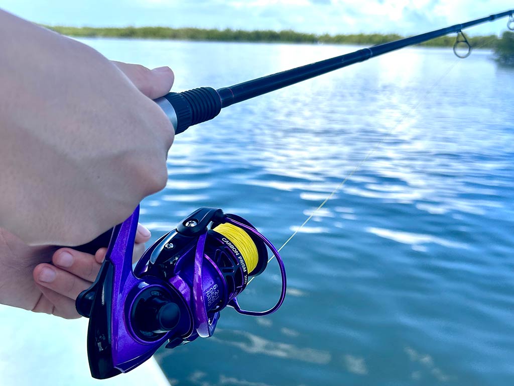 A closeup of a small fishing reel being held along with a rod on inshore saltwater fishing grounds, with calm water visible out of focus in the background