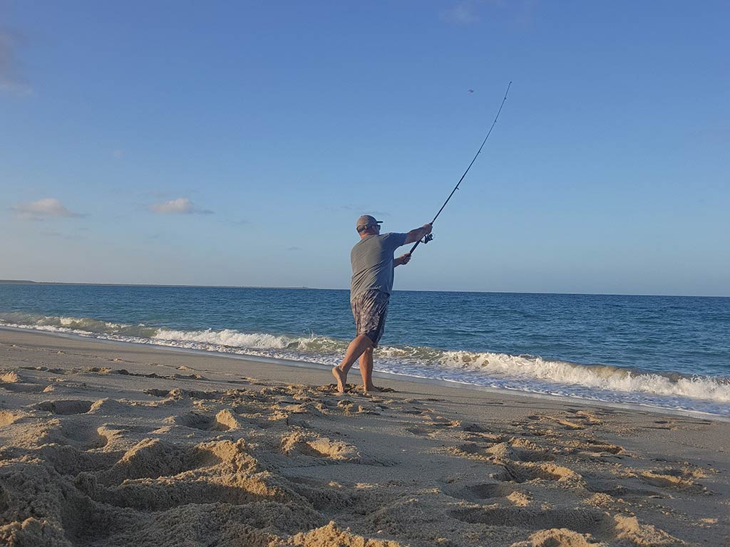 A view from behind of a man casting from the surf with a long rod into the ocean in Mexico at sunset on a bright day