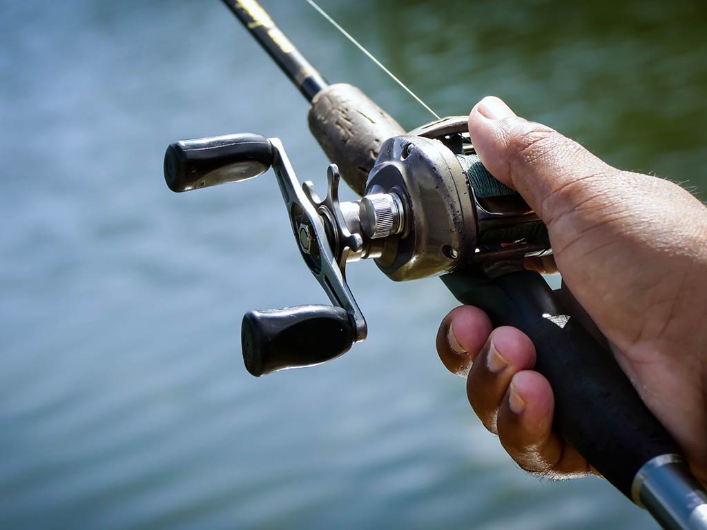 A closeup of a hand holding a fishing rod next to a baitcaster reel, with a lake or river out of focus in the background