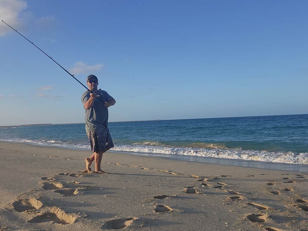 A man looks towards the camera as he prepares to cast his fishing rod from the surf into the sea in Mexico, with footprints in the sand visible in the foreground