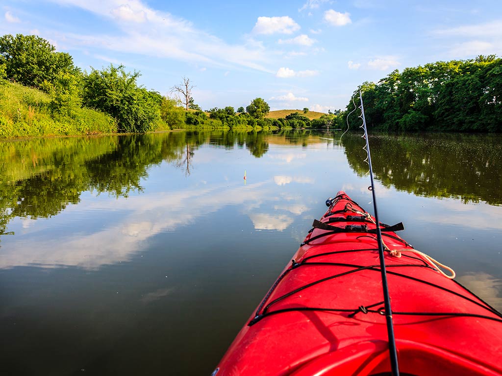 A view along a river in Kentucky from a red kayak with a fishing rod hanging over the front of the boat on a clear day