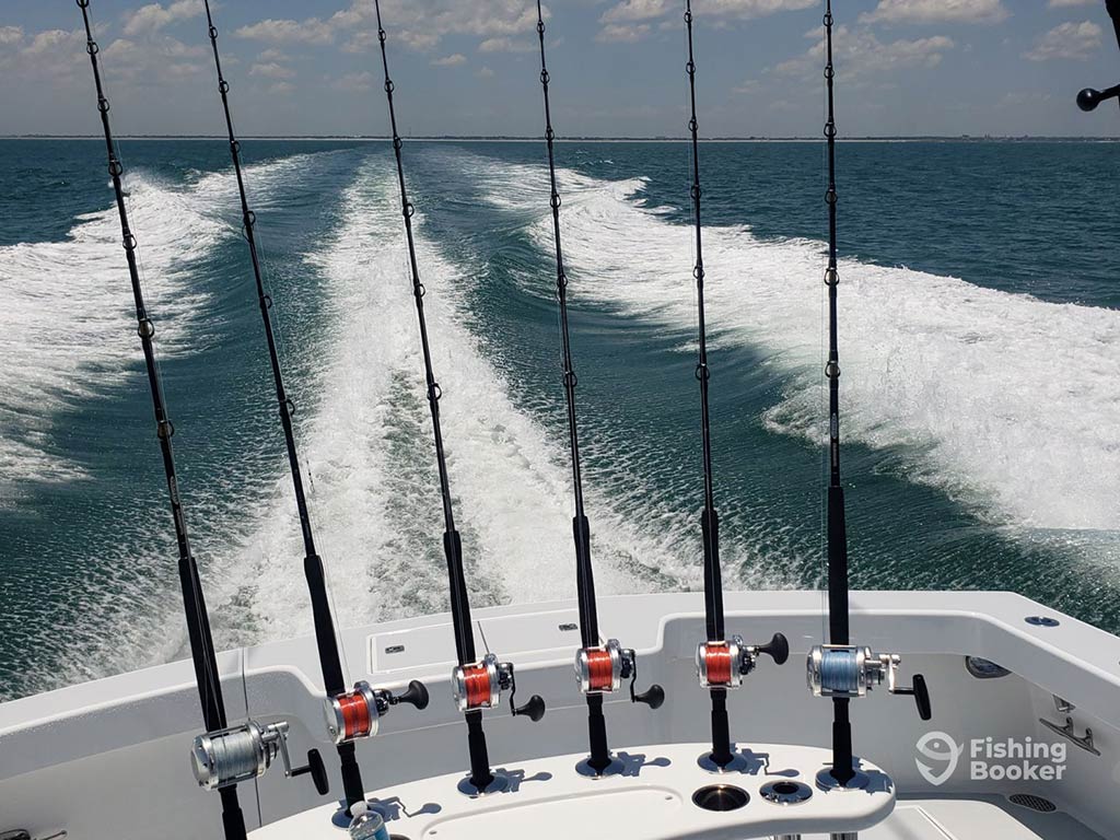 A view out across the back of a fishing boat with six trolling rods lined up and the wake of the boat visible in the water behind