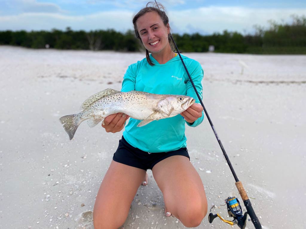 A woman kneeling on a beach and holding a Speckled Trout with a St.Croix Mojo inshore fishing rod next to her