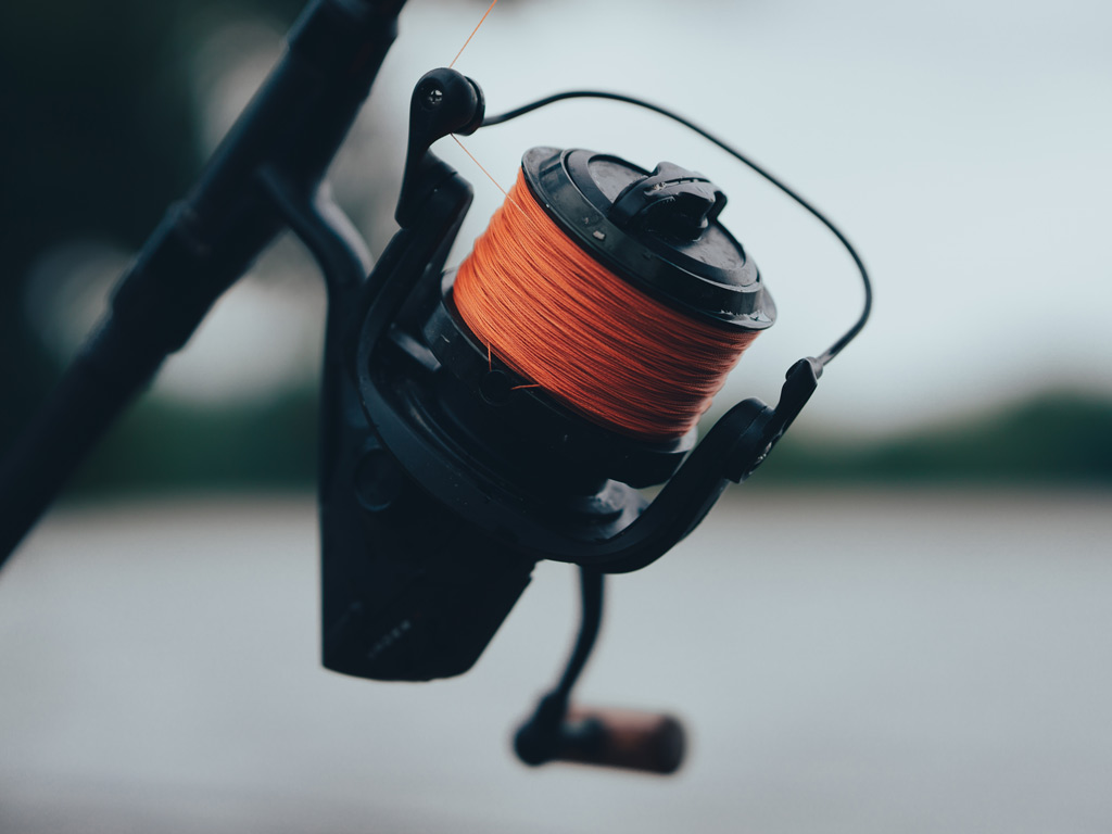 A closeup of a spinning reel spooled with orange braided line, typically a combo that'd be fairly expensive as beginner fishing gear, with blurred background behind the reel.