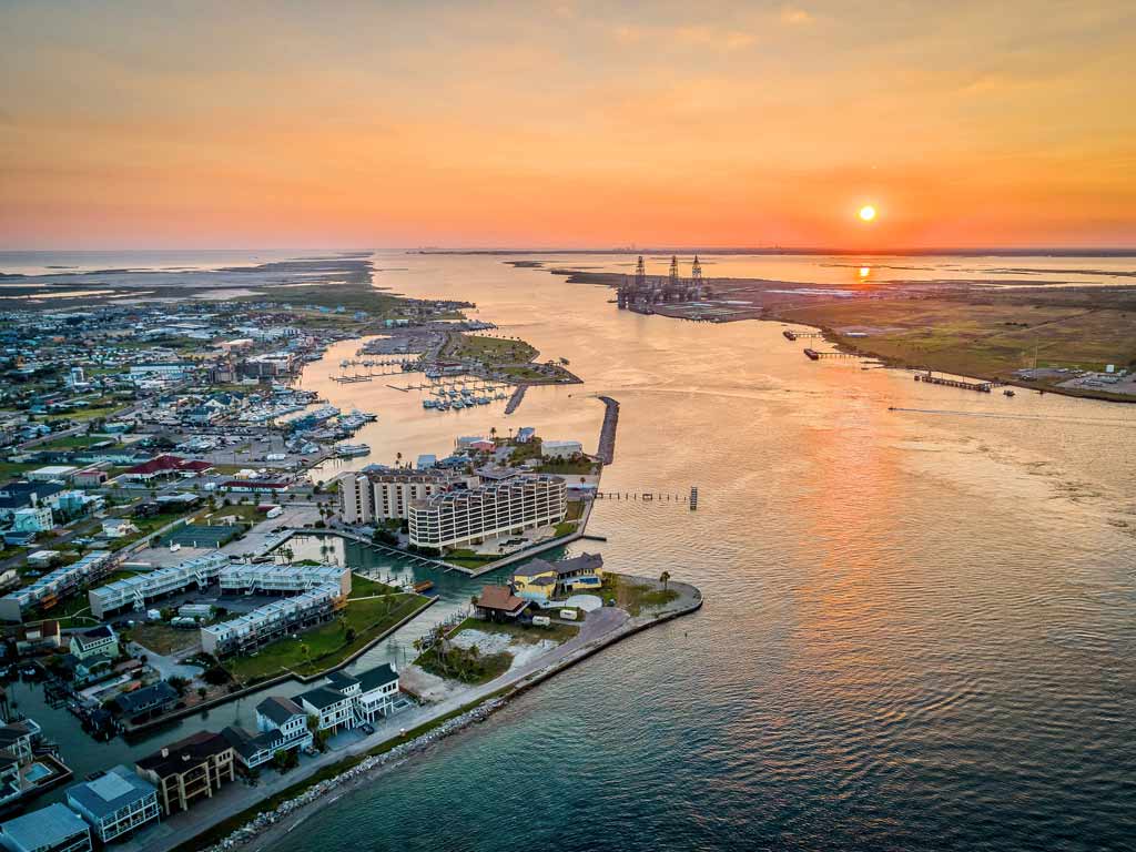 A sunset view of the Intracoastal Waterway in Port Aransas, with the city buildings and the harbor visible on the left side of the photo while the channel separates it from the other shore on the right.