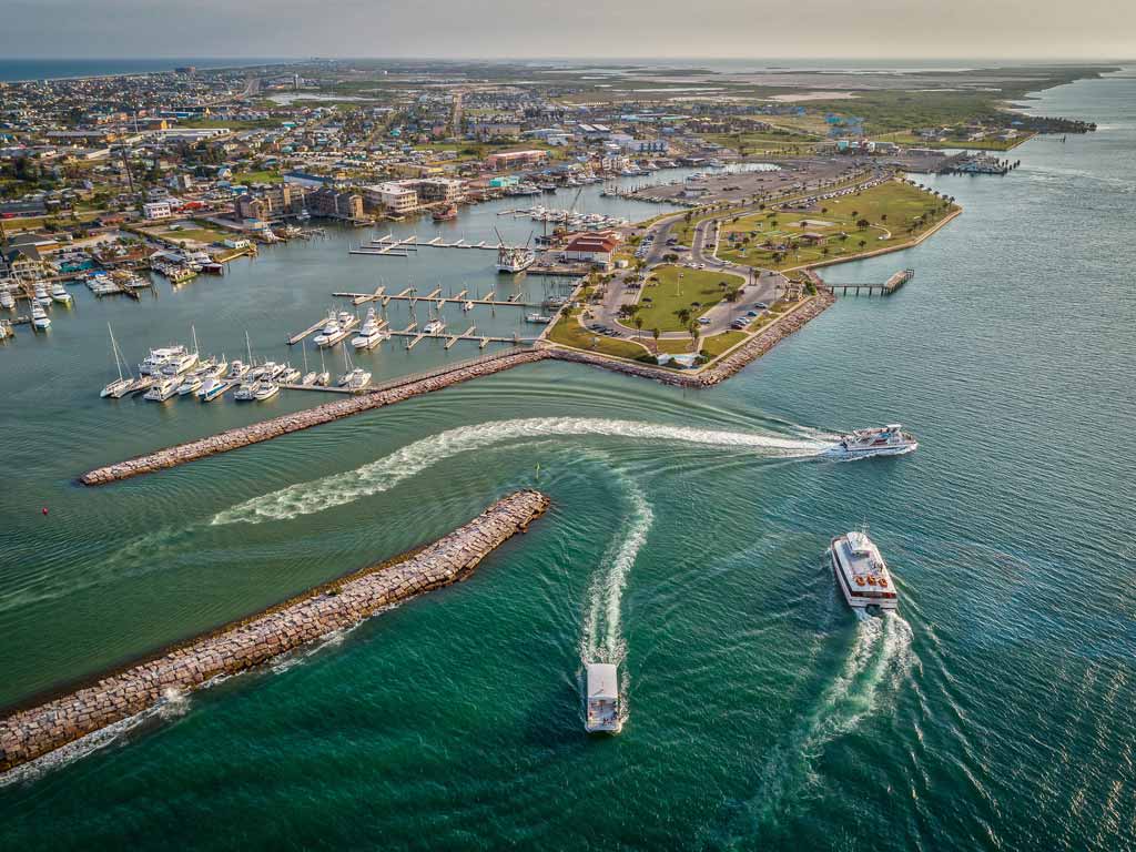 An aerial view of the Port Aransas Municipal Harbor and multiple boats going in and out of it, with the rest of the city visible in the distance.