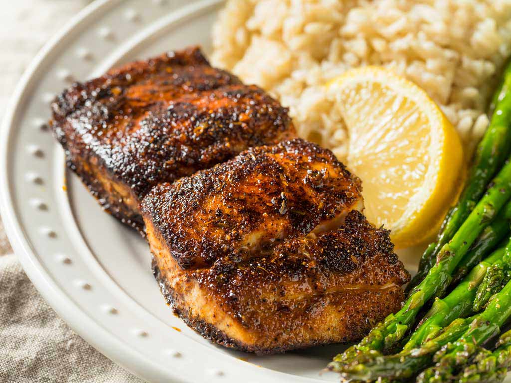 A closeup shot of two grilled Mahi Mahi fillets on a white plate, along with a side of rice, asparagus, and a sliced lemon.