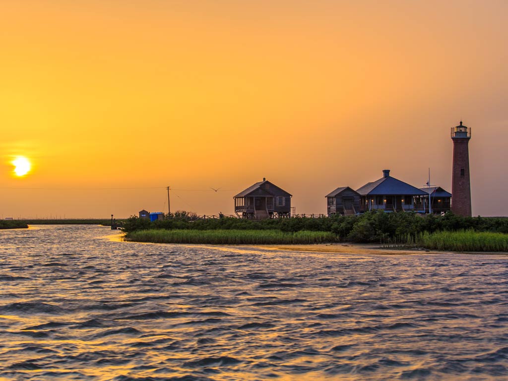 A view of the Lydia Ann Lighthouse in Aransas Pass at sunset, with orange skies reflecting and darkening the water surface in focus, and the lighthouse visible to the right of the image.