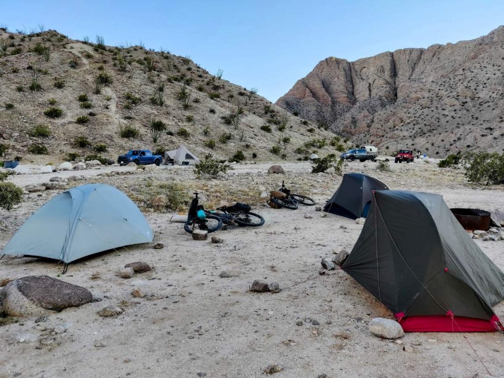 Three bikepacking tents at a desert campground with canyon walls and several trucks and campers in background.