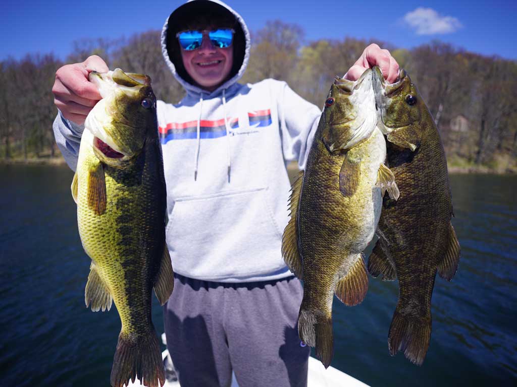 A man in sunglasses and with his hood up over his head standing on a fishing boat and holding one Largemouth Bass and two Smallmouths by their mouths on a sunny day