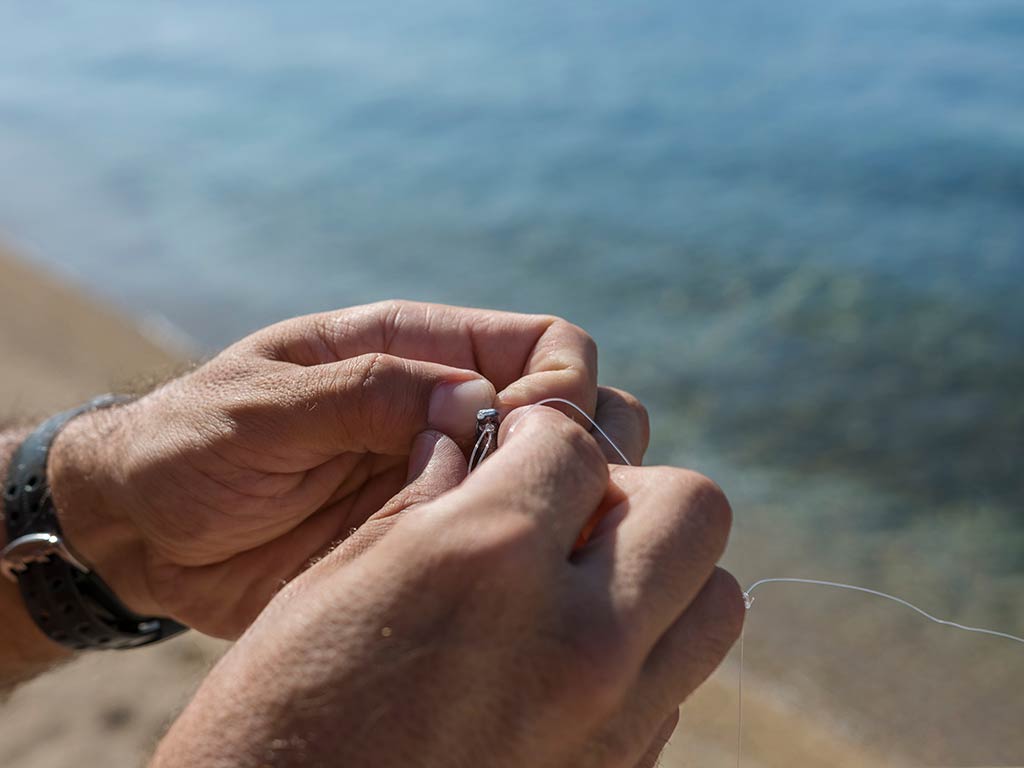 A closeup of two hands tying a small knot on a fishing line next to the water's edge on a bright day