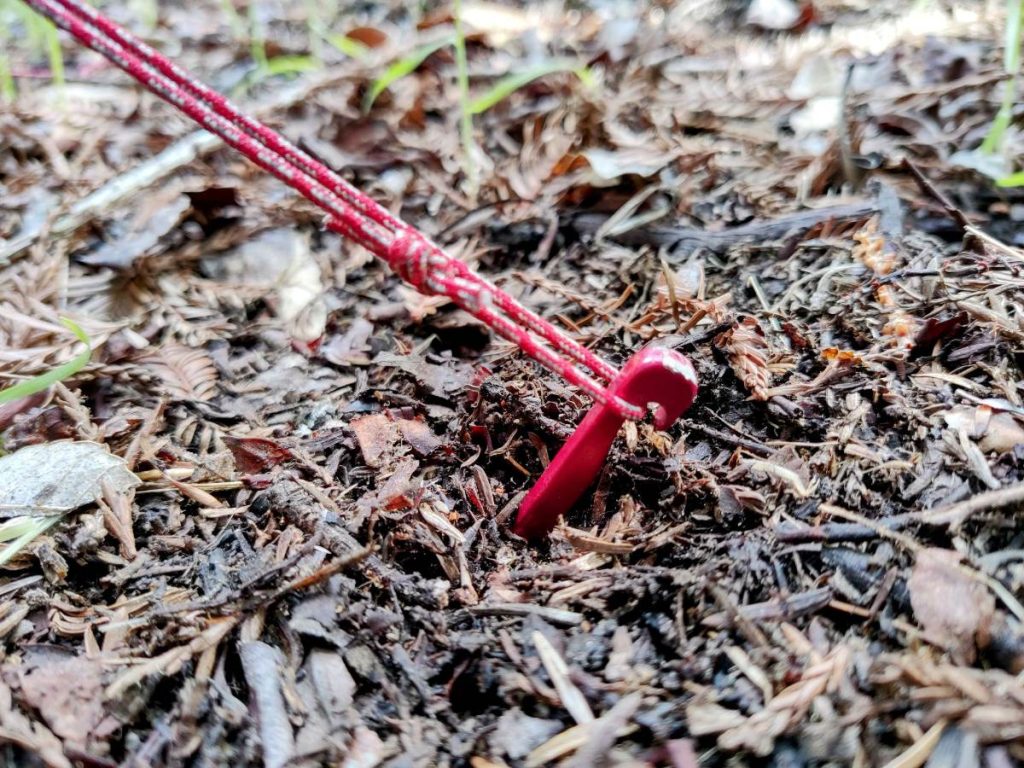 Close up of red tent stake pounded into ground at an angle, and a guy line pulling at right angle to the stake
