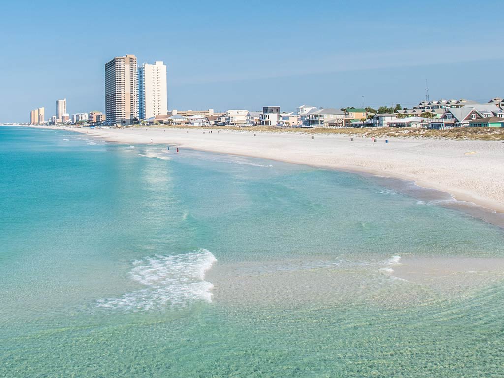 A view of the beachfront in Panama City Beach on a sunny day, with turquoise shallow waters and white sand in the forefront and city buildings in the distance.