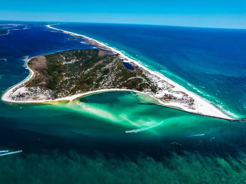 An aerial view of Saint Andrews State Park, with the waters surrounding the park changing colors from dark blue to turquoise, while multiple boats are seen going in and out of the nearby inlet.